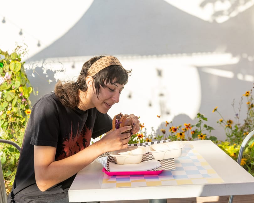 A person in a black shirt and leopard print headband eats a burger at an outdoor table.