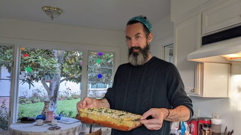 A man in a kitchen holds a glass baking dish filled with a layered casserole.