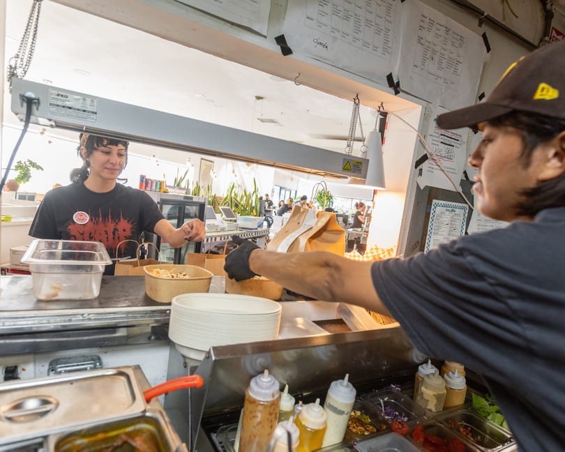 Two restaurant workers preparing food and packaging orders at a service counter with sauces and ingredients.