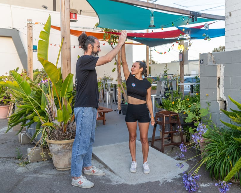 Two people high-fiving outdoors under colorful shade sails, surrounded by lush plants and string lights.