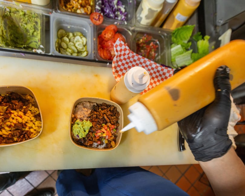 Chef in black gloves squeezing sauce onto a bowl of food with various toppings in containers.