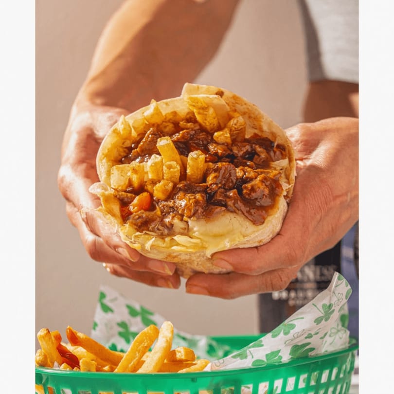 Hands holding a loaded pita with meat, sauce, and french fries, with a basket of fries below.