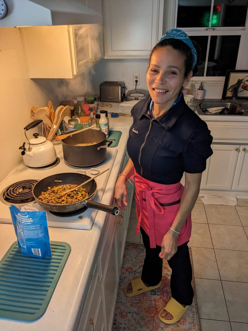 A smiling woman in a blue top and pink apron cooks in a kitchen with steaming pots on the stove.
