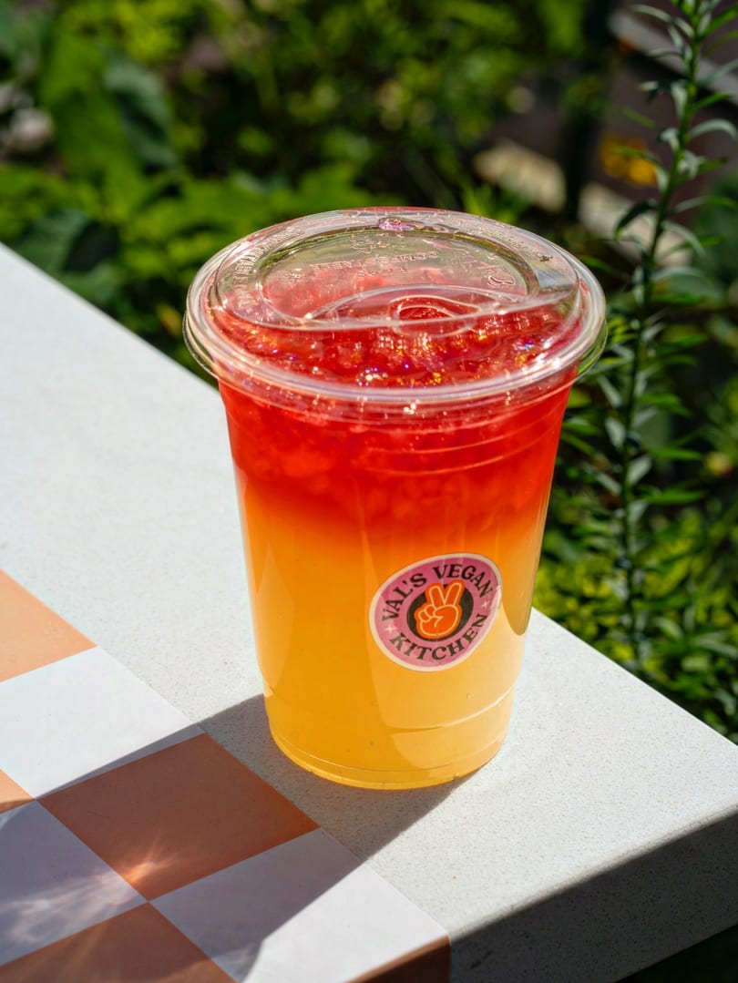 A Val's Vegan Kitchen drink, red on top and yellow on the bottom, sits on a tiled surface.