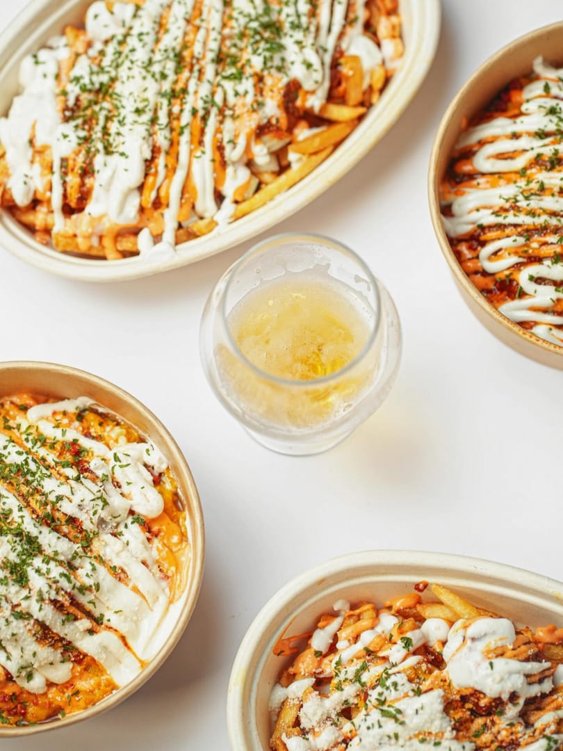 Four bowls of loaded fries with various sauces and herbs, surrounding a glass of beer.