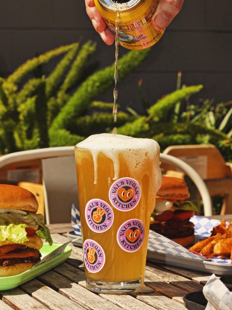 Beer being poured into a Val's Vegan Kitchen glass, with burgers and fries on a wooden table.
