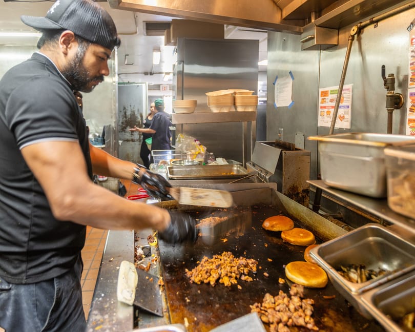 Chef in a black cap and shirt cooking meat and buns on a griddle in a busy kitchen.