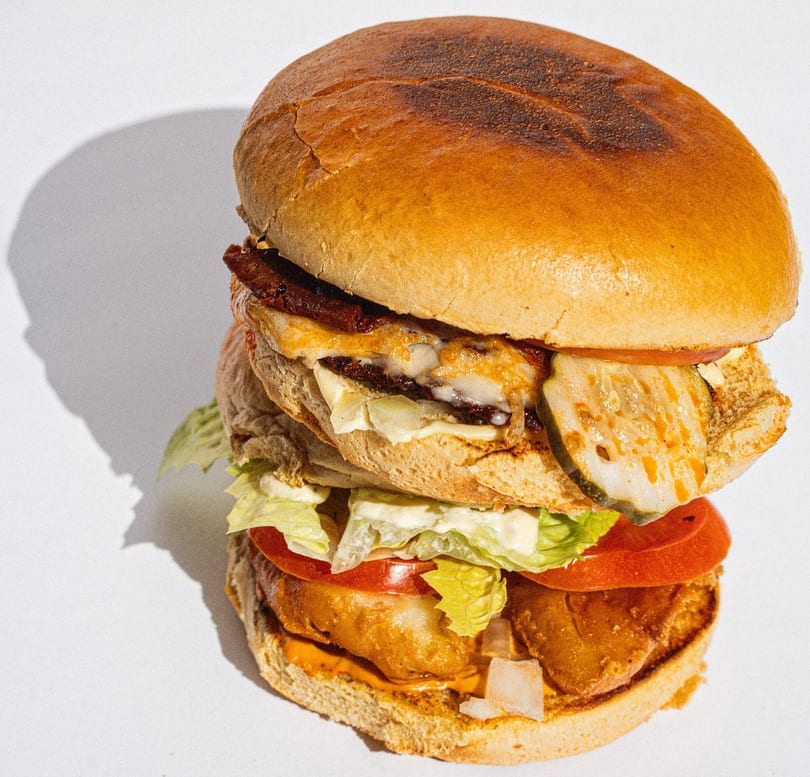 Tall double-decker burger with cheese, bacon, pickles, lettuce, tomato, and sauce on a white background.