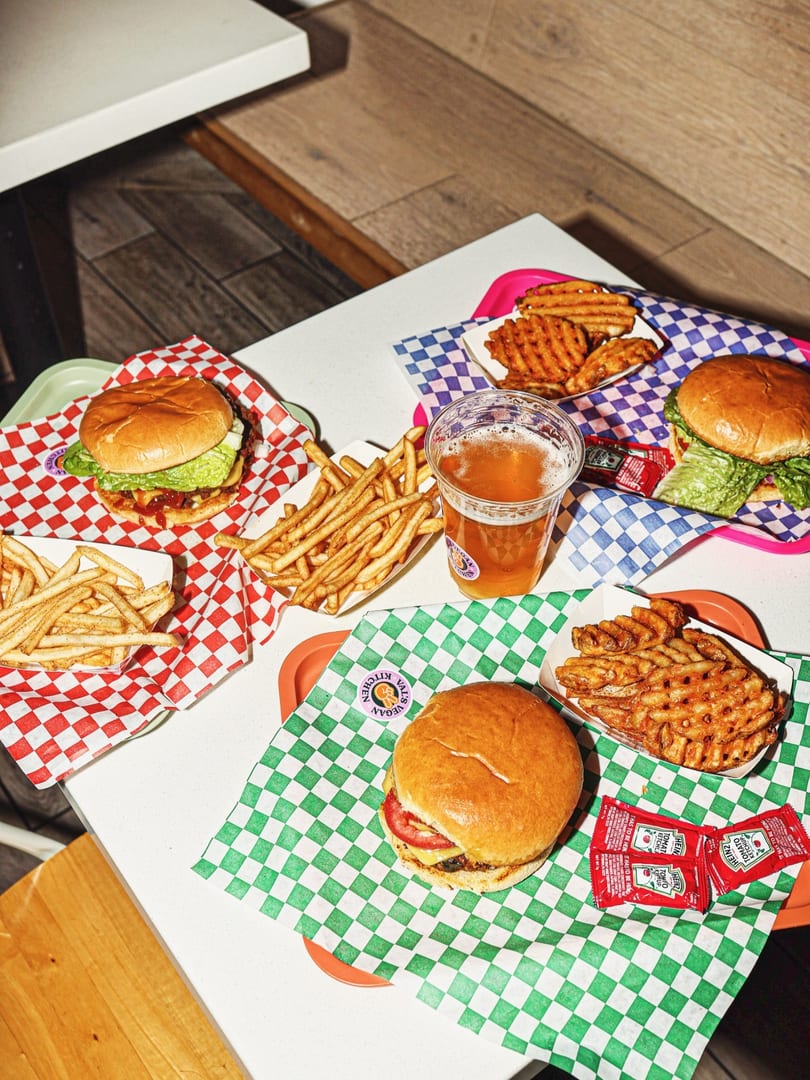 Three burgers, two with fries, one with waffle fries, and a beer on a white table.