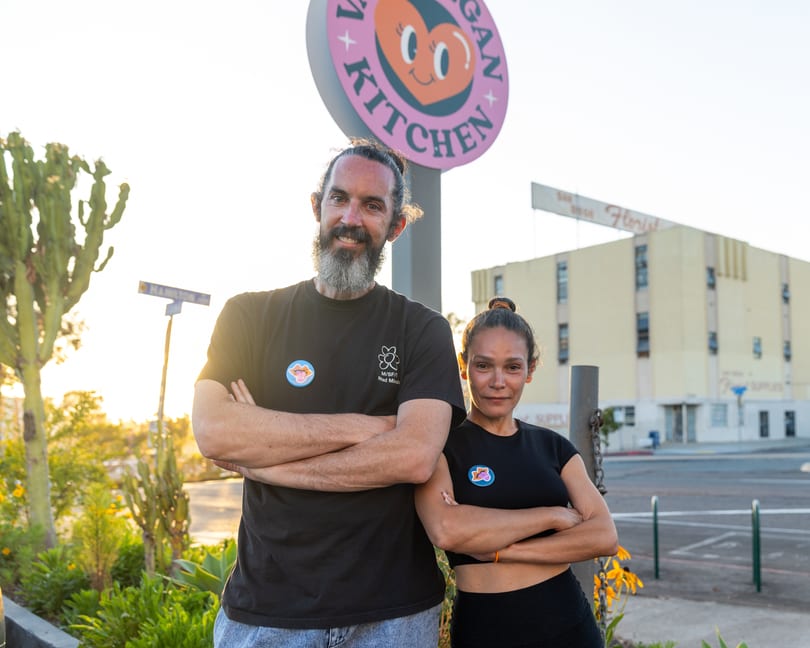 Two smiling people, a man and a woman, stand with crossed arms in front of a "Vegan Kitchen" sign.