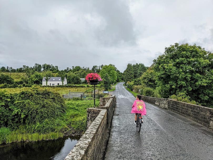 Person in a pink Nirvana jacket cycling over a stone bridge in a lush green rural landscape.